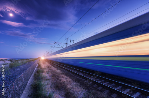 High speed passenger train in motion on the railroad at summer night. Moving blurred modern commuter train at dusk. Railway station and purple sky with moon and clouds. Industrial landscape. Transport