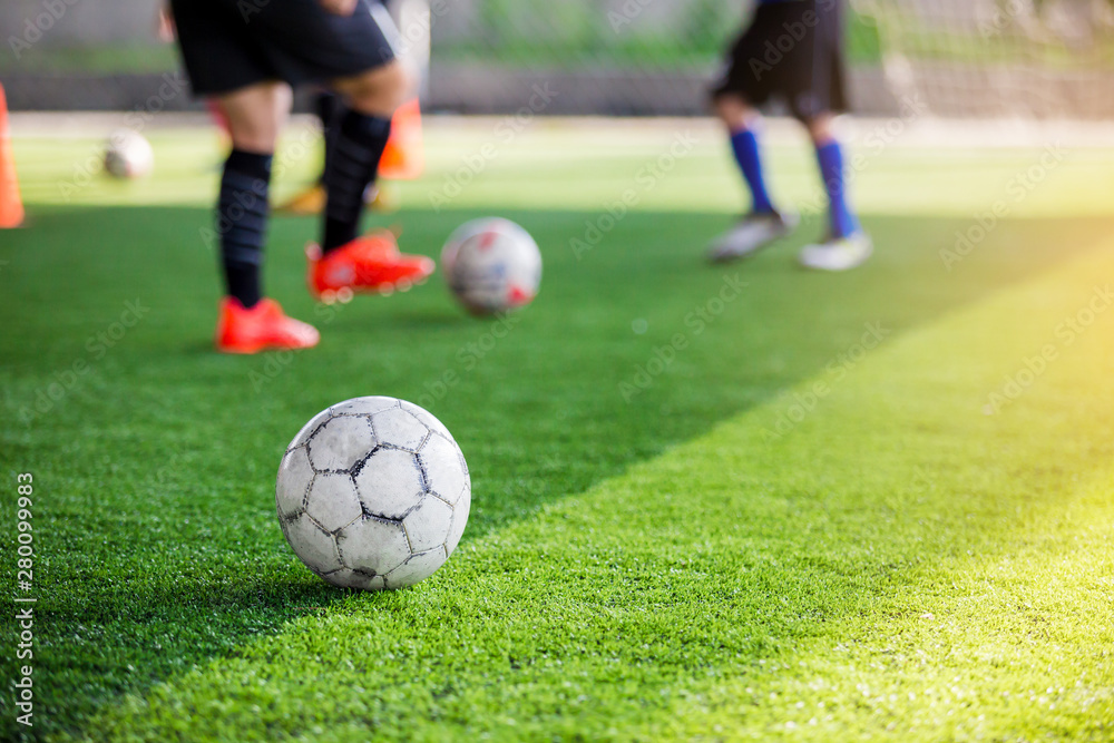 football between marker cones on green artificial turf with blurry soccer team training, blurry kid soccer player jogging between marker cones and control ball with soccer equipment in soccer academy.