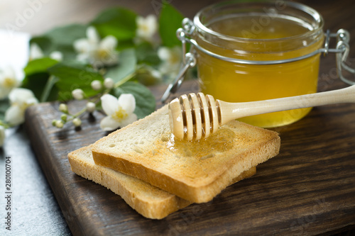 wooden spoon with honey is lying on a piece of bread near a glass jar of honey