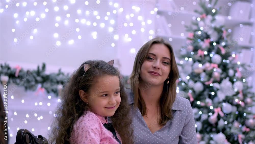 Young attractive mother and daughter sitting hugging looking at the camera smiling against the background of Christmas lights and Christmas tree. Young attractive mother and daughter sitting hugging looking at the camera smiling against the background of Christmas lights and Christmas tree.