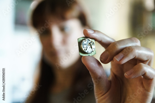 Portrait of woman watchmaker. Girl with blurred face holds clock in hand. Clockwork in fingers. Gears in disassembled watches on blurred background.