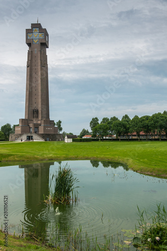 Diksmuide  Flanders  Belgium -  June 19  2019  IJzertoren  tallest peace monument of WW 1 against gray blue cloudscape. On wall saying No More War. Reflection in pond. Some green foliage and lawn. 