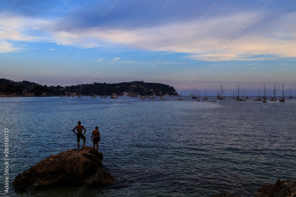 View of the beach, two bathers on a rock, bay of Beaulieu sur Mer, Saint Jean Cap Ferrat at sunset