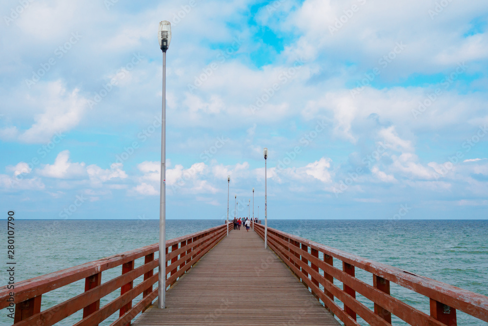 Sea piercing with a pier equipped with wooden railings and lanterns. Blue sky with white cumulus clouds.