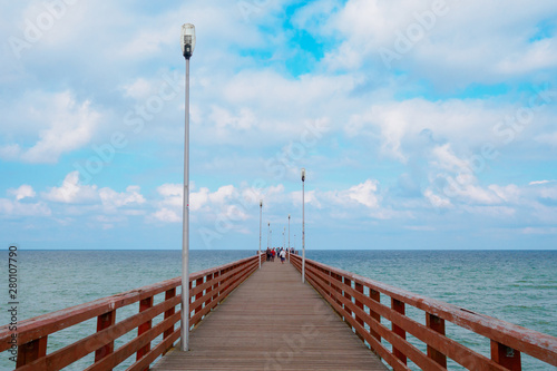 Sea piercing with a pier equipped with wooden railings and lanterns. Blue sky with white cumulus clouds.