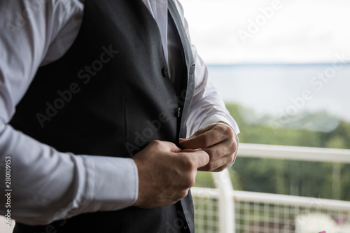 the groom fastens the vest on the terrace against the backdrop of nature and the lake