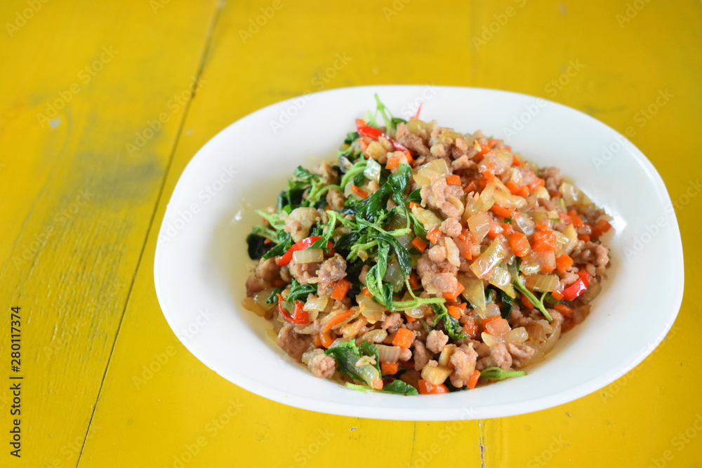 Spicy Thai food, stir-fried pork basil in a white dish placed on a yellow wooden table. Spicy Thai food, stir-fried pork basil in a white dish placed on a yellow wooden table.