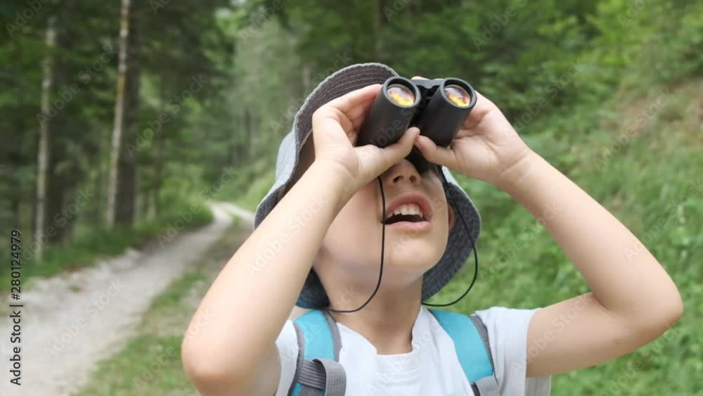 slow motion, a caucasian child watches with binoculars, wears a blue hat and behind him there is a path in the woods, front view slow motion, a caucasian child watches with binoculars, wears a blue hat and behind him there is a path in the woods, front view