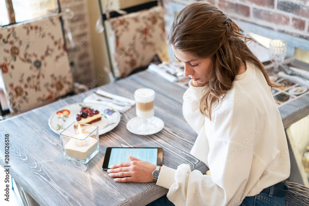 beautiful girl during breakfast in cafe reads e-book. Work on tablet during break. Freelance. Creative person. Latte coffee, cheesecake.