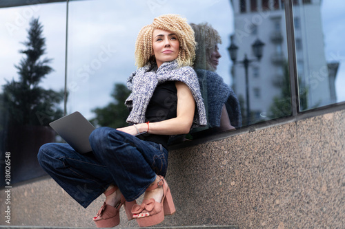 young hipster girl, afro hair, curls working with laptop on street near glass showcase. Freelance on open space.