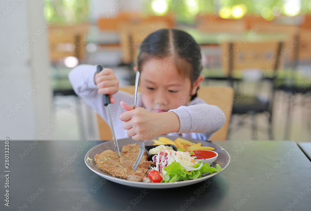 Asian child girl eating Pork steak and vegetable salad on the table with holding knife and fork. Children having breakfast. Asian child girl eating Pork steak and vegetable salad on the table with holding knife and fork. Children having breakfast.