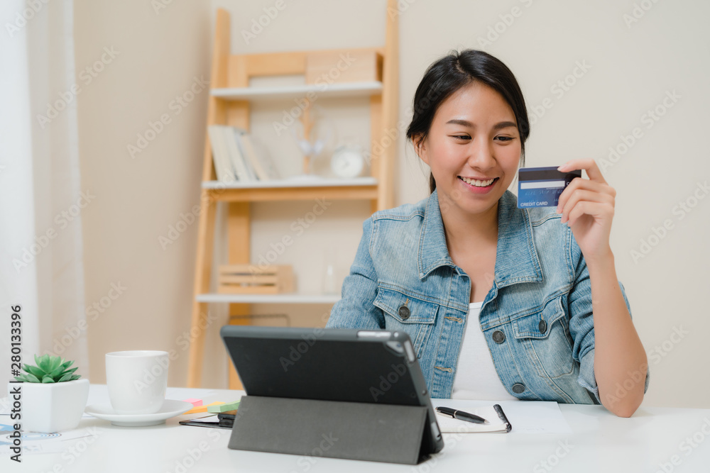 Beautiful smart business Asian woman using tablet buying online shopping by credit card while wear smart casual sitting on desk in living room at home. Lifestyle woman working at home concept. Beautiful smart business Asian woman using tablet buying online shopping by credit card while wear smart casual sitting on desk in living room at home. Lifestyle woman working at home concept.