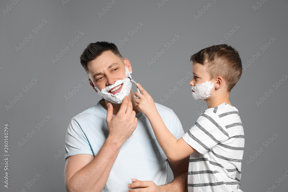 Son shaving his dad with razor on grey background Son shaving his dad with razor on grey background