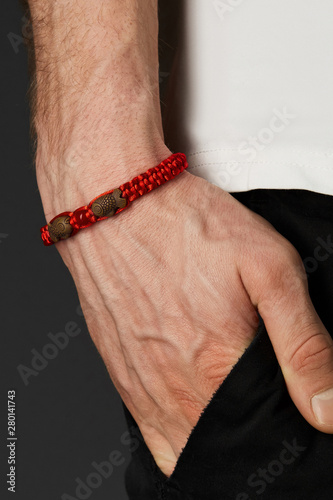 Cropped closeup shot of a man with a bracelet on hшы wrist. The red bracelet is weaving with brown wood insets. The fashion model dressed in jeans and checkered shirt is posing on the black background