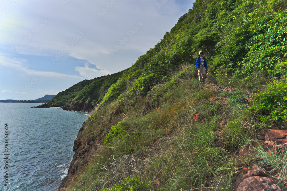 Female adventurer walking on the rocky gravel trail path out of the forest on the cliff nearby the sea in the summer afternoon with beautiful clouds and sky
