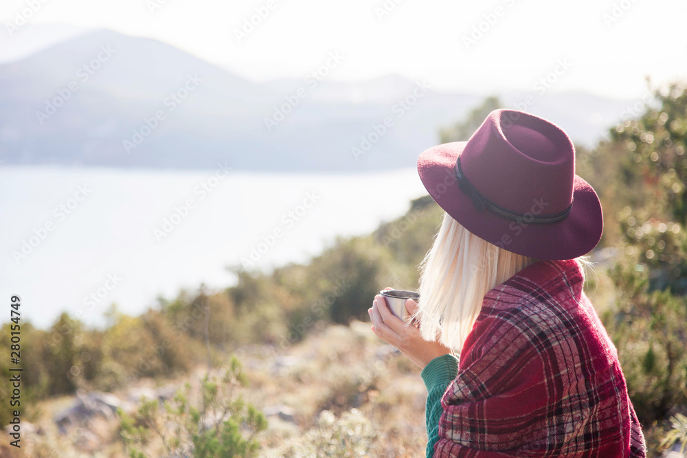Woman traveler in mountains above autumn sea. Cozy fall picnic with coffee on beach. Girl in warm plaid and hat drinks hot steamy tea from mug. Wellbeing, relaxation at nature, traveling concept.