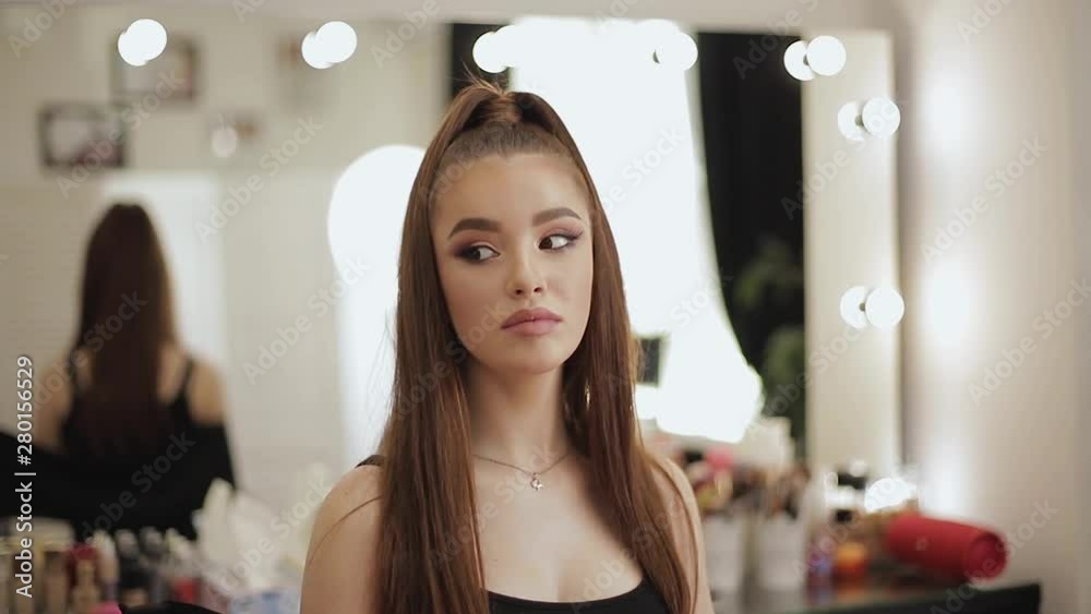 Young beautiful model looking in the mirror after makeup. Attractive young woman sitting in the beauty studio. Background of mirror in beauty salon Young beautiful model looking in the mirror after makeup. Attractive young woman sitting in the beauty studio. Background of mirror in beauty salon
