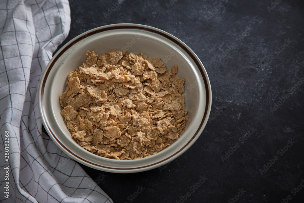 Wheat bran breakfast cereal with no milk in a bowl. Black background with homespun napkin. Wheat bran breakfast cereal with no milk in a bowl. Black background with homespun napkin.