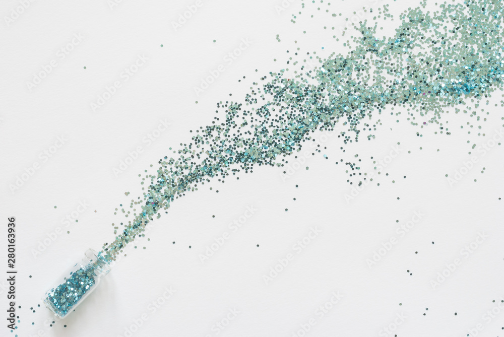 Turquoise blue glitter confetti spill out of a small glass jar diagonally on a white paper background. Turquoise blue glitter confetti spill out of a small glass jar diagonally on a white paper background.