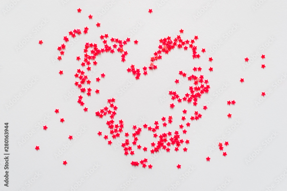 Red glitter stars are laid out in the shape of a heart on a white paper background. Close up flat lay.