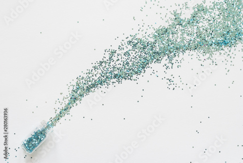Turquoise blue glitter confetti spill out of a small glass jar diagonally on a white paper background.