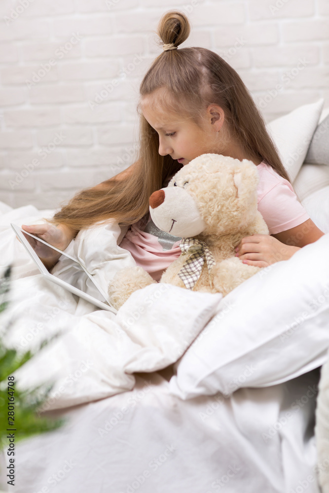 cute little child girl with teddy bear lies in bed uses digital tablet. child playing on tablet pc. cute little child girl with teddy bear lies in bed uses digital tablet. child playing on tablet pc.