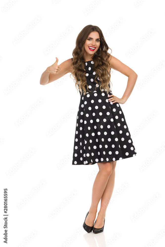 Beautiful Young Woman In Black Cocktail Dress In Polka Dots And High Heels Is Showing Thumb Up. Beautiful Young Woman In Black Cocktail Dress In Polka Dots And High Heels Is Showing Thumb Up.