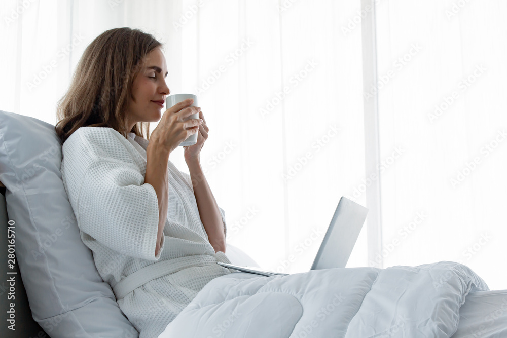 Young caucasian woman in white pajamas with white holding a cup of coffee and drink and find information from the laptop on a bed. Young caucasian woman in white pajamas with white holding a cup of coffee and drink and find information from the laptop on a bed.