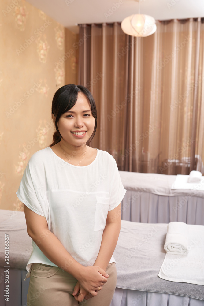 Portrait of Asian massage therapist standing near the massage table and smiling at camera she waiting for her client in spa salon Portrait of Asian massage therapist standing near the massage table and smiling at camera she waiting for her client in spa salon