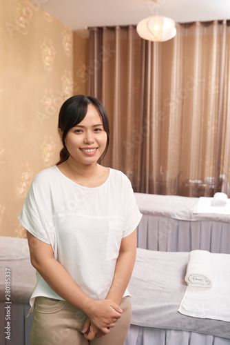 Portrait of Asian massage therapist standing near the massage table and smiling at camera she waiting for her client in spa salon