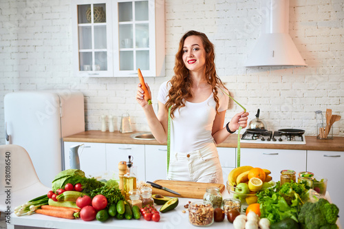 Young happy woman holding carrot in the beautiful kitchen with green fresh ingredients indoors. Healthy food and Dieting concept. Loosing Weight