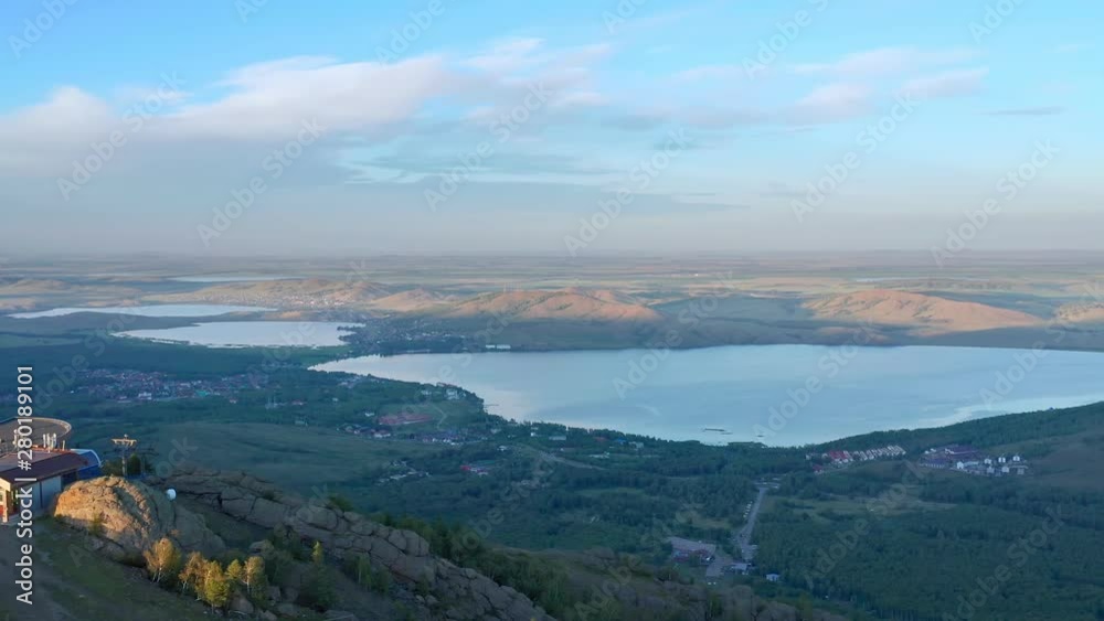 The lateral shot over the mountain peak. At the top of the mountain is a building. In the background is a lake and mountains. Near the lake is a village. Evening hours. The lateral shot over the mountain peak. At the top of the mountain is a building. In the background is a lake and mountains. Near the lake is a village. Evening hours.