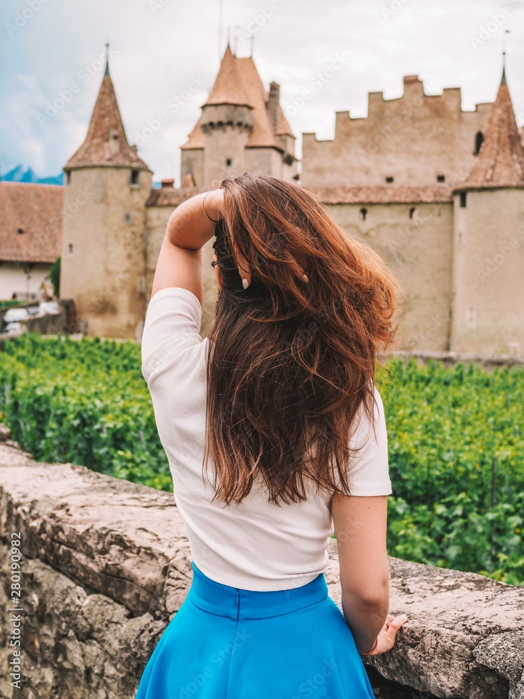 A girl with long hair in a blue skirt stands with a view of Aigle Castle and the Vineyards in the province near the city of Montreux in Switzerland, the mountains