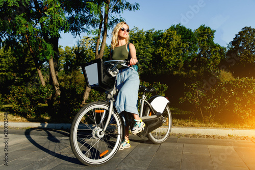 Full-lenght photo of blonde in sunglasses looking at side in long denim skirt standing on bike next to green bushes in city