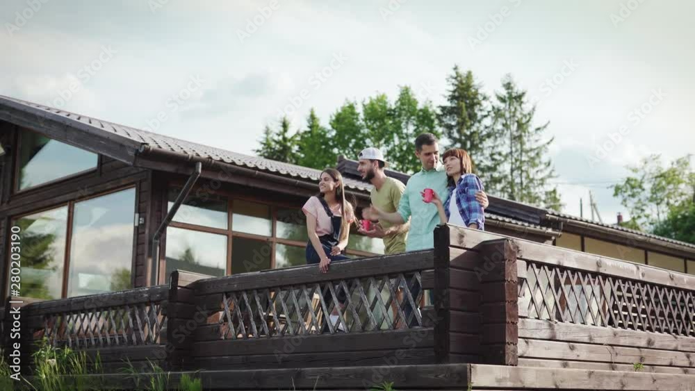 Camera moving away from four young friends, two couples, standing at railing of wooden country house terrace, looking at view and talking, panning shot