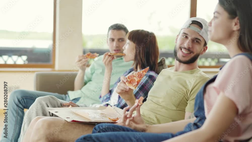 Side view tracking shot of four diverse young friends sitting on sofa, eating delicious pizza and talking. Focus on charismatic man flirting with girlfriend Side view tracking shot of four diverse young friends sitting on sofa, eating delicious pizza and talking. Focus on charismatic man flirting with girlfriend