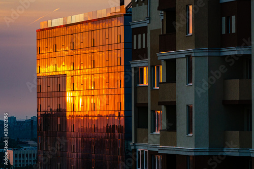 Sunset among multi-storey urban buildings. Sun's rays are reflected in orange in glass windows of tall building. Close-up. Voronezh, Russia, June, 2019