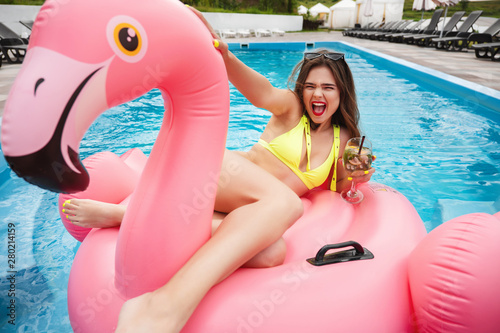 young brunette gorgeous woman with perfect body wearing yellow trendy sexy swimsuit suntanning in the hotel pool drinking tasty cocktail on pink inflatable flamingo © studioprodakshn