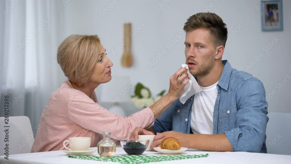 Mother wiping irritated adult sons mouth during having tea at home, care Mother wiping irritated adult sons mouth during having tea at home, care