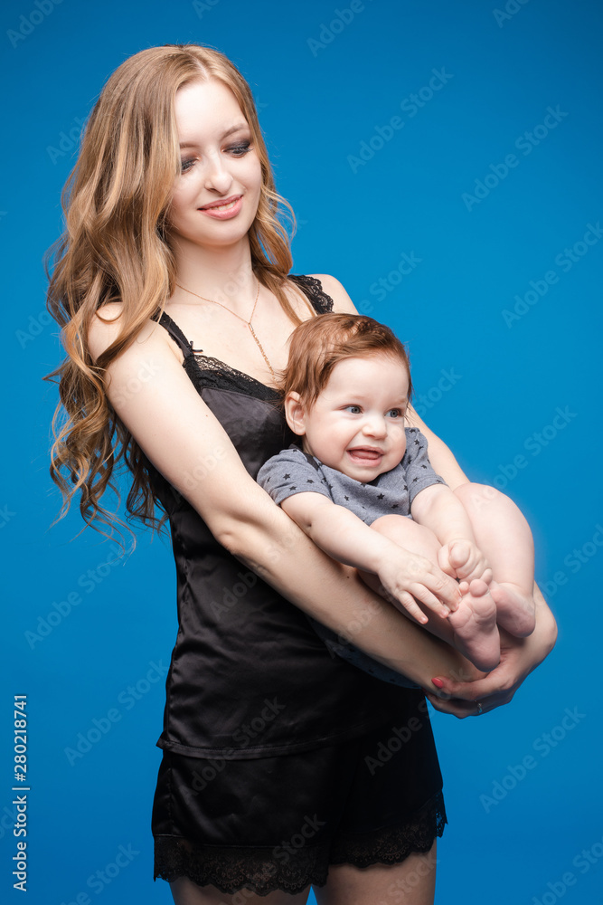 Front view of positive little baby laughing with mother in studio. Beautiful woman in black pajamas keeping cute daughter on hands. looking at camera and posing on blue isolated background.
