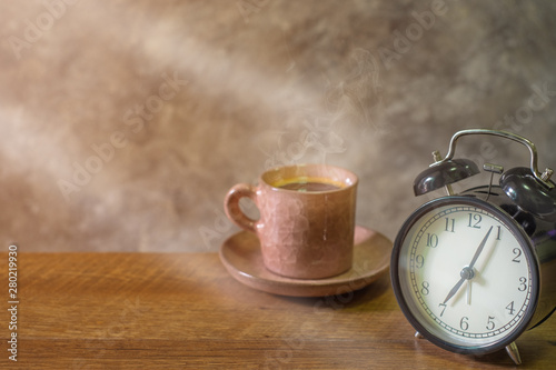 Close-up of retro alarm black clock vintage style with cup coffee pastel pink old on wooden table. Time for Wake up early. light morning through the window. background cement wall in home.