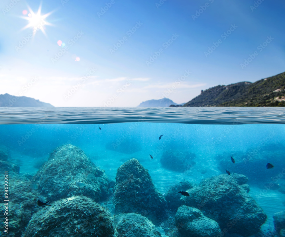Blue sunny sky, an island in the ocean and underwater sandy rocky sea view.