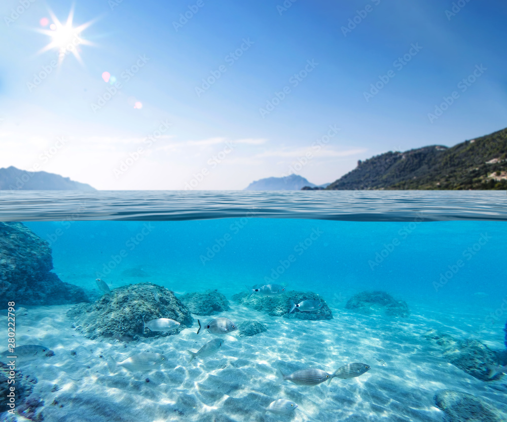 Blue sunny sky, an island in the ocean and underwater sandy rocky sea view.