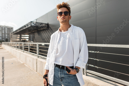 Photography American young handsome man fashion model in white stylish shirt in blue vintage jeans in trendy sunglasses is posing on the street in the sunny day