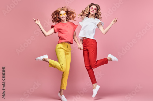 Two fashionable girl jump Smiling in colorful outfit on pink. Beautiful easy-going woman in red yellow pants, Stylish curly hair having fun. Joyful funny slim sisters friends, happy fashion concept
