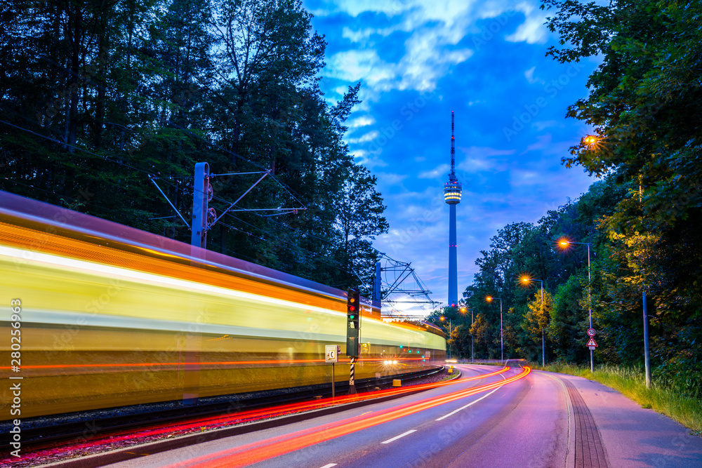 Germany, Illuminated tramway passing by street of traffic next to television tower of stuttgart city in green forest nature landscape in magical twilight mood after sunset