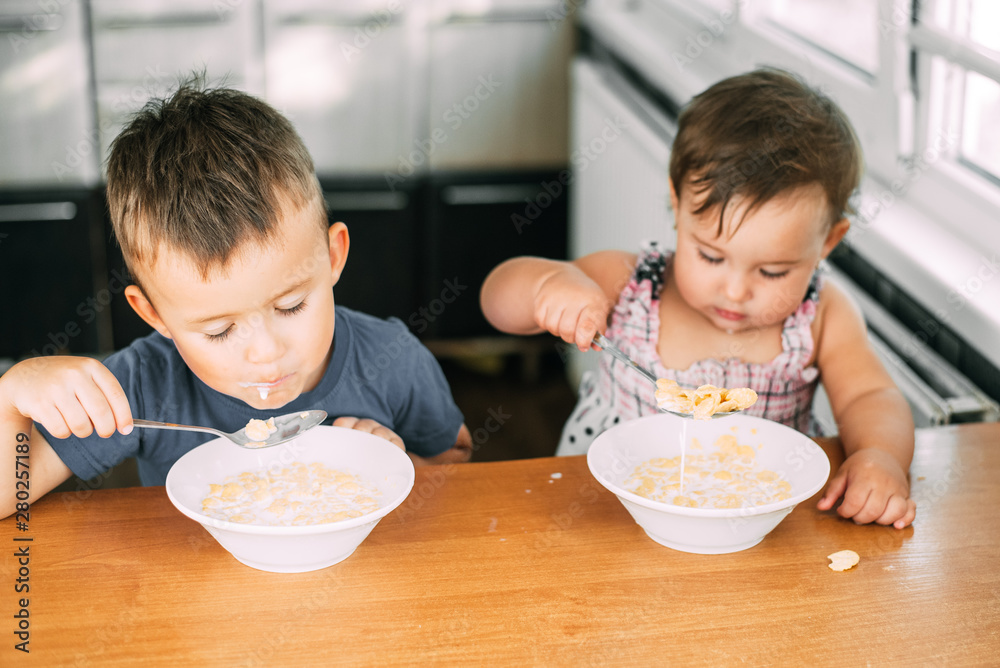 Brother and sister eat cornflakes with milk in the kitchen during the day very fun and cute, dirty and funny