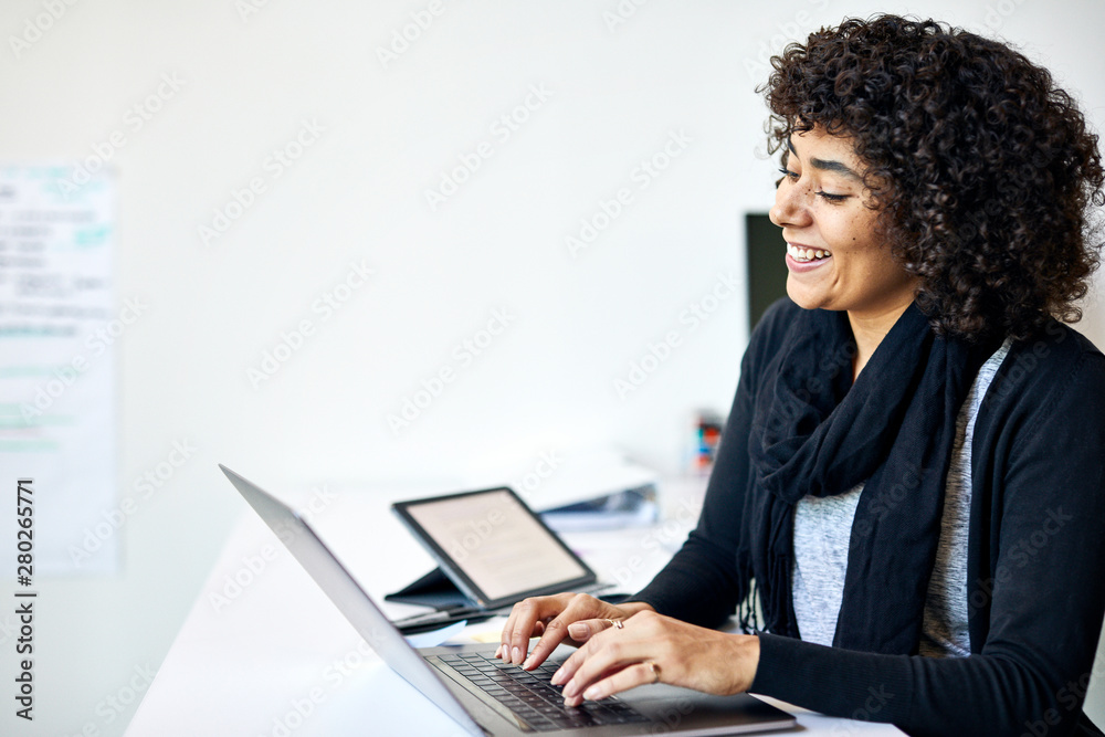 Smiling businesswoman with curly hair using laptop computer on desk against wall in office