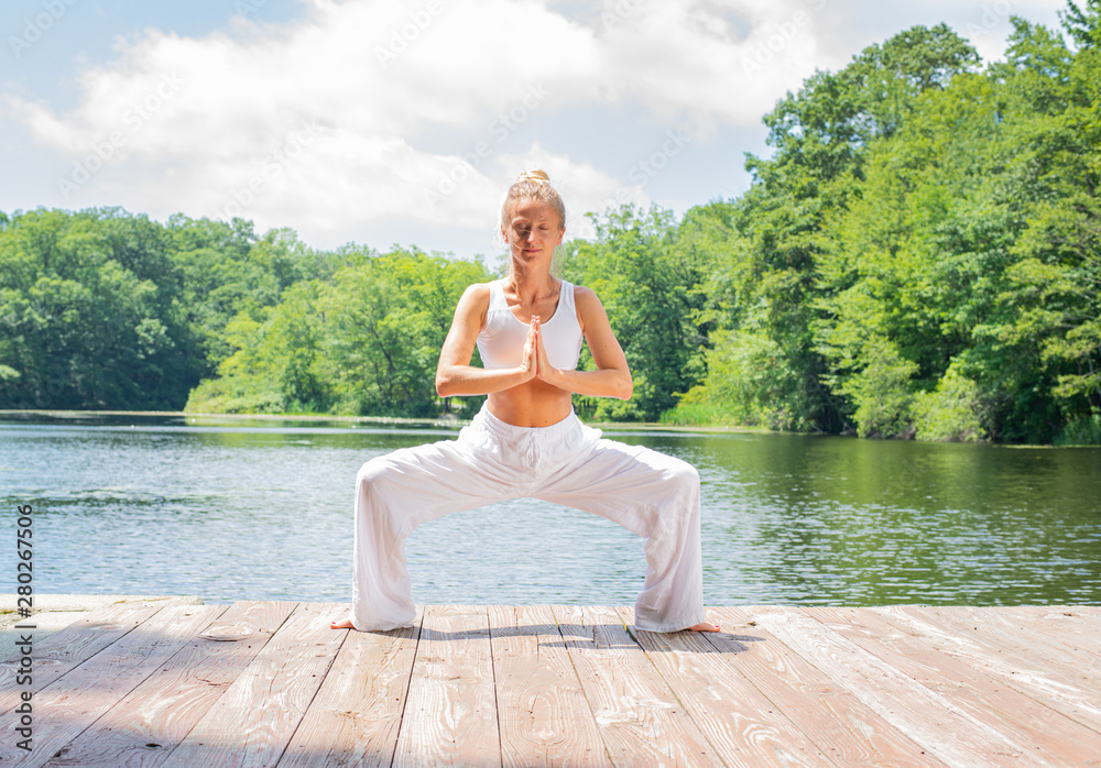 Attractive woman is practicing yoga, doing Stupasana exercise, standing in Goddess pose near lake.