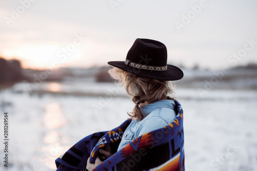 woman stood in a field looking towards the sunset in a hat & blanket photo